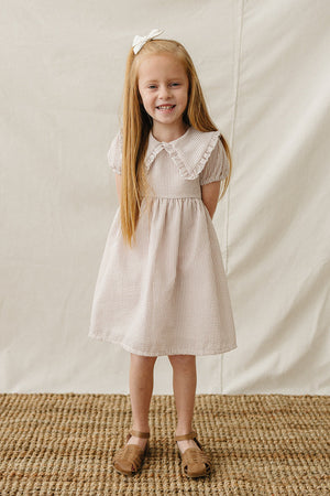 A smiling girl wears a taupe striped cotton dress with a Peter Pan collar, standing on a textured woven rug.