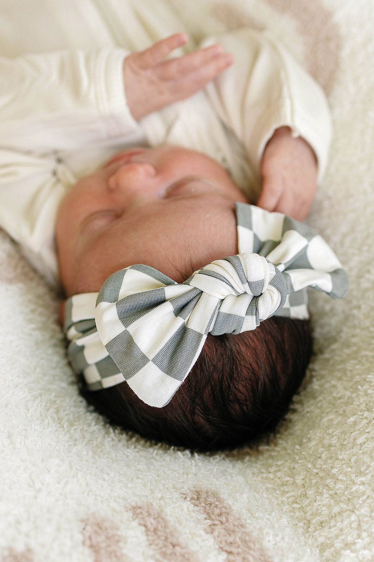 A baby wearing a light green checkered head wrap, lying on a soft, textured blanket.