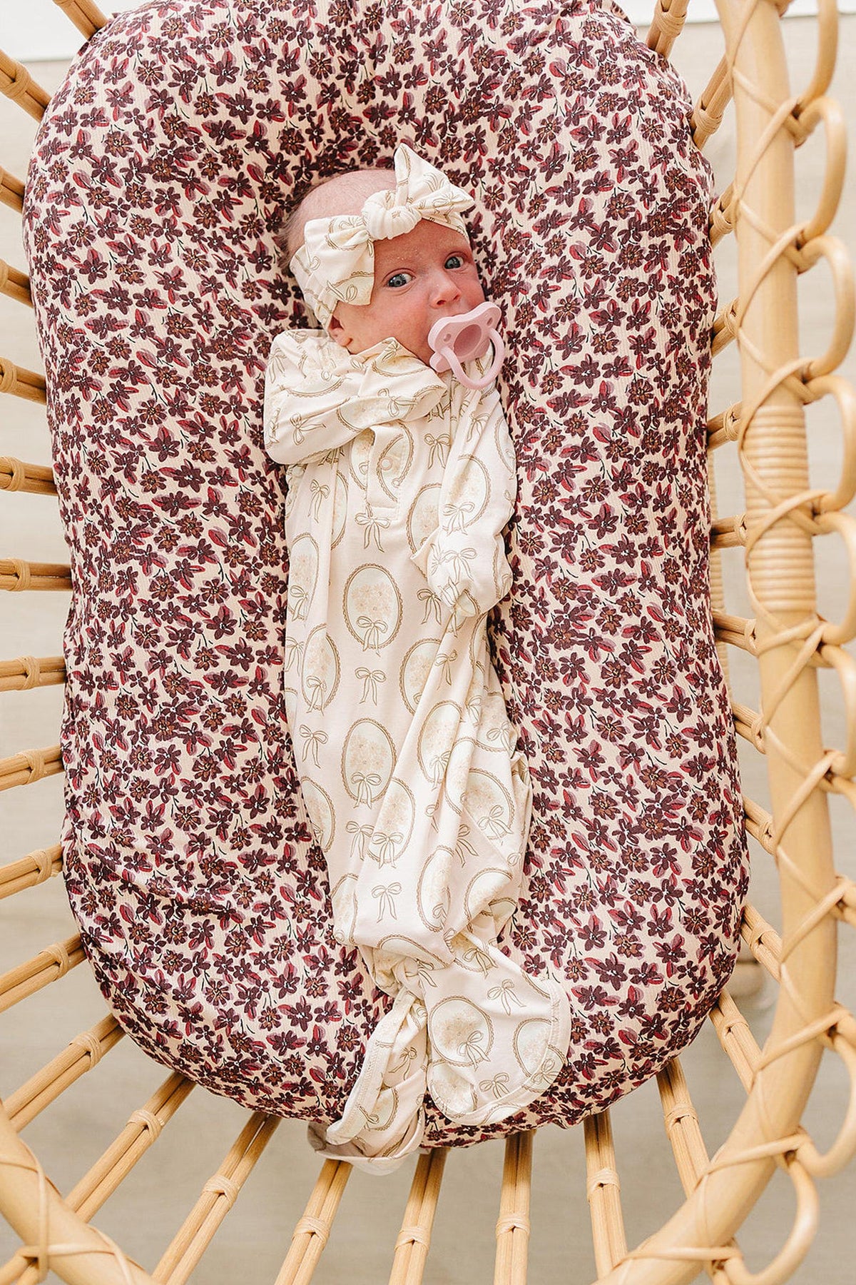 A newborn baby is swaddled in a soft, cream-colored bamboo gown featuring a vintage floral print, lying comfortably in a cozy, patterned bassinet with a matching headband.