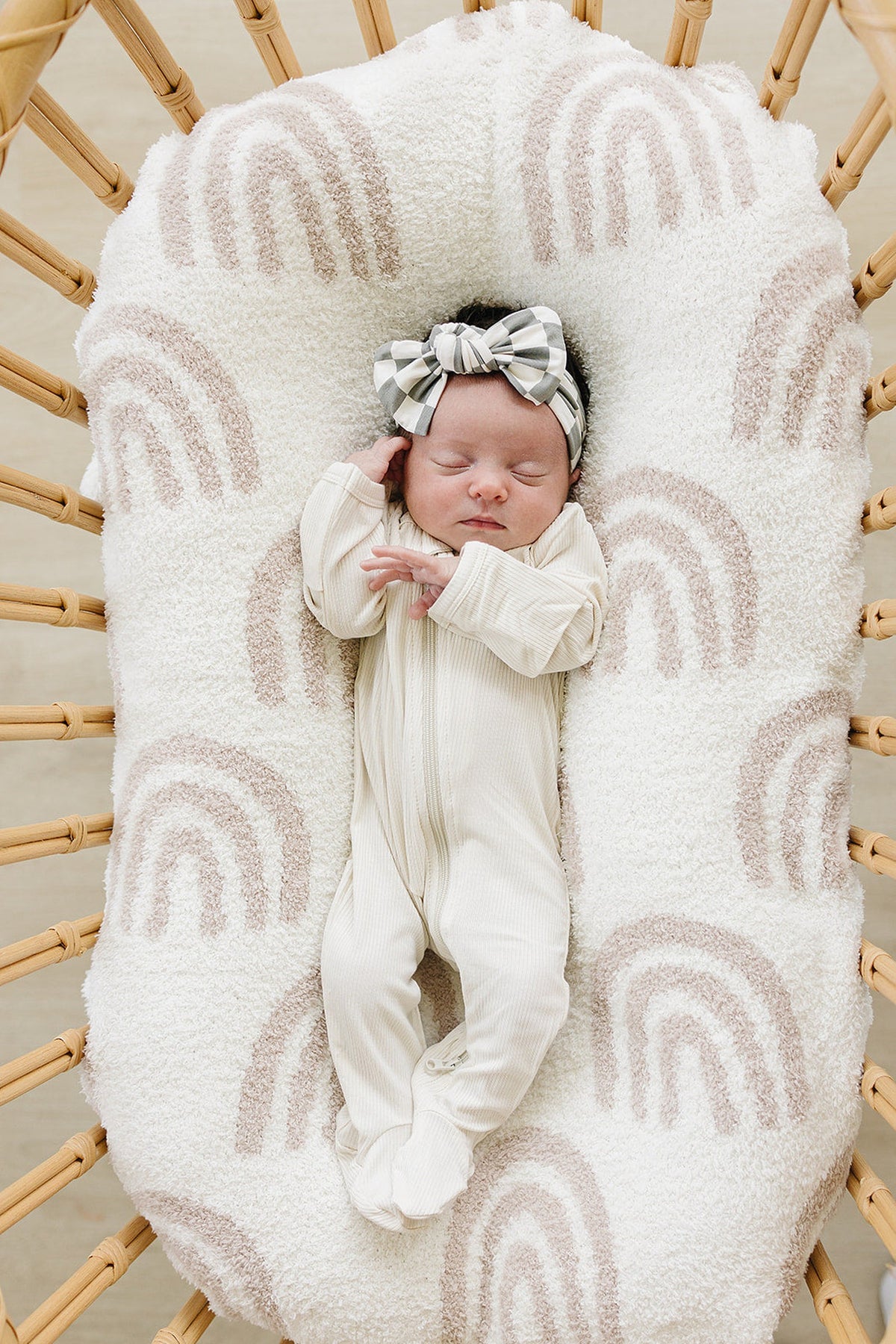 A sleeping baby wearing a light green checkered head wrap and cream outfit lies on a rainbow-patterned cushion.