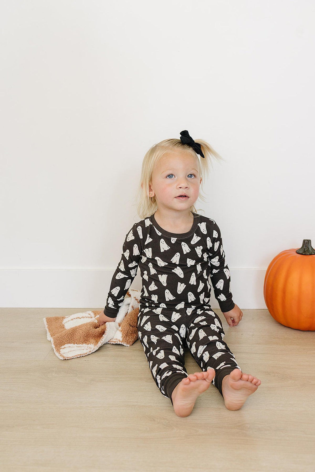 A child in ghost-print pajamas sits on the floor with a pumpkin and a blanket nearby.