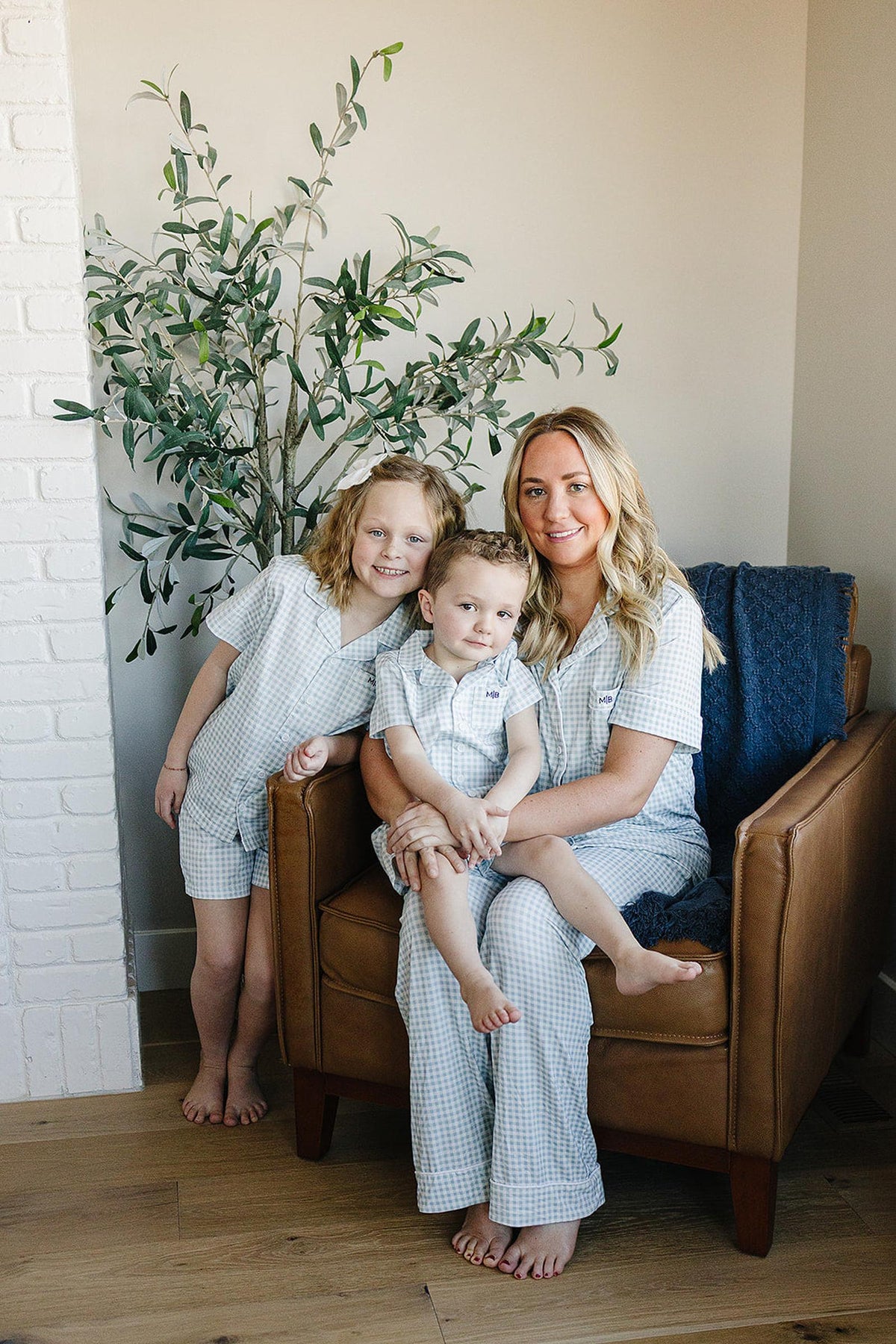 A woman and two children wearing matching light blue gingham pajamas sit on a brown armchair indoors.