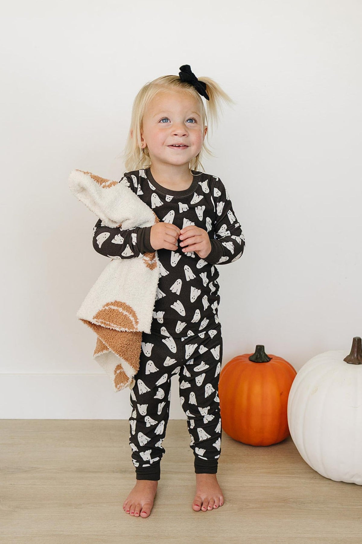 A child in ghost-print pajamas holds a blanket, standing beside orange and white pumpkins.