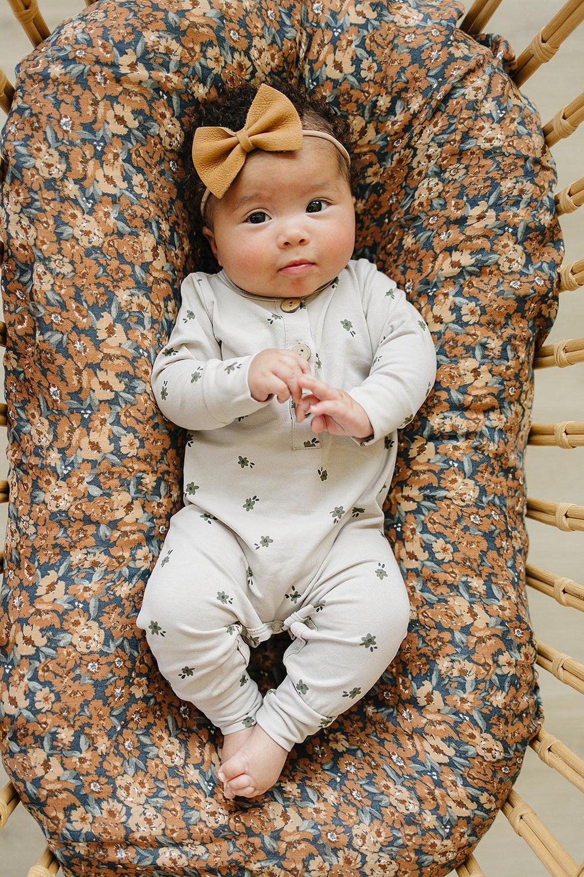 Baby wearing a light-colored floral romper with button-up front, lying on a floral-patterned cushion, and a brown bow.