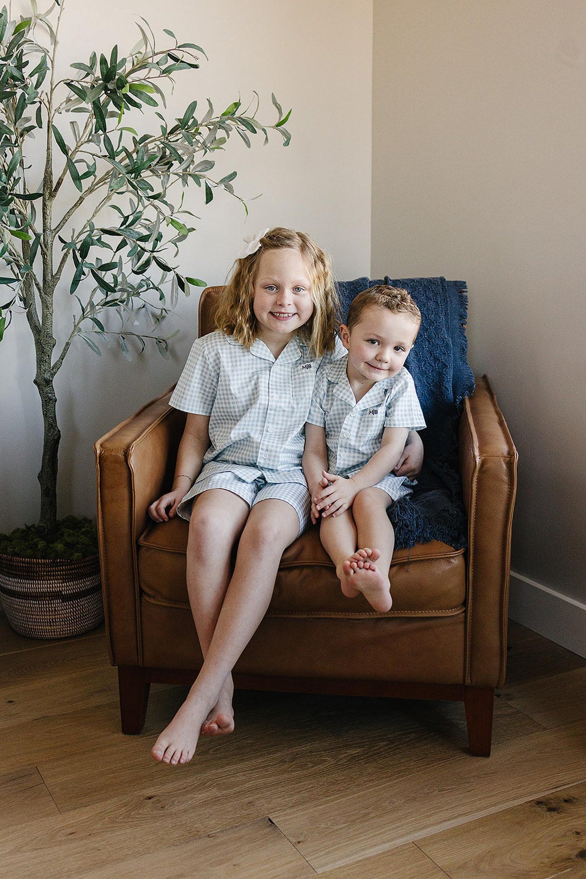 Two children sit on a brown armchair, wearing matching light blue gingham pajamas. A potted plant is nearby.
