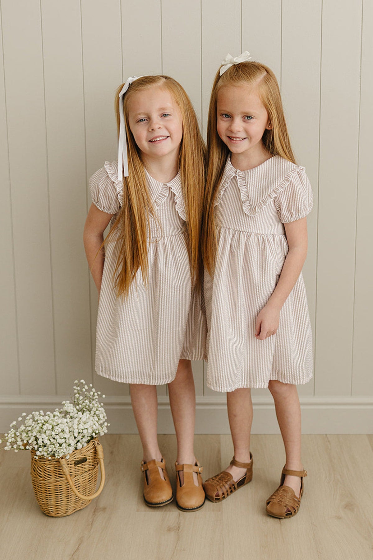 Two girls wear taupe striped dresses with Peter Pan collars, standing in front of a wooden wall, holding a basket.