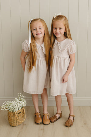 Two girls wear taupe striped dresses with Peter Pan collars, standing in front of a wooden wall, holding a basket.