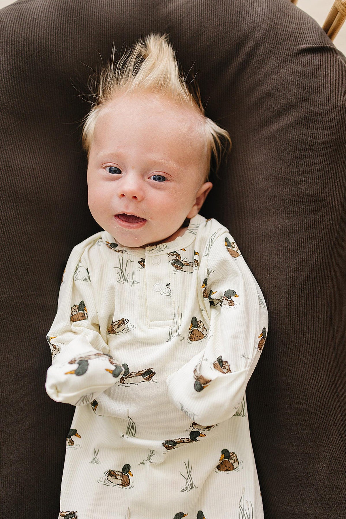 A smiling baby with spiky hair is wearing a cream-colored ribbed bamboo gown adorned with duck motifs, resting against a dark brown fabric background. The gown is soft and eco-friendly.