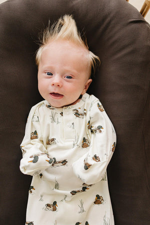 A smiling baby with spiky hair is wearing a cream-colored ribbed bamboo gown adorned with duck motifs, resting against a dark brown fabric background. The gown is soft and eco-friendly.