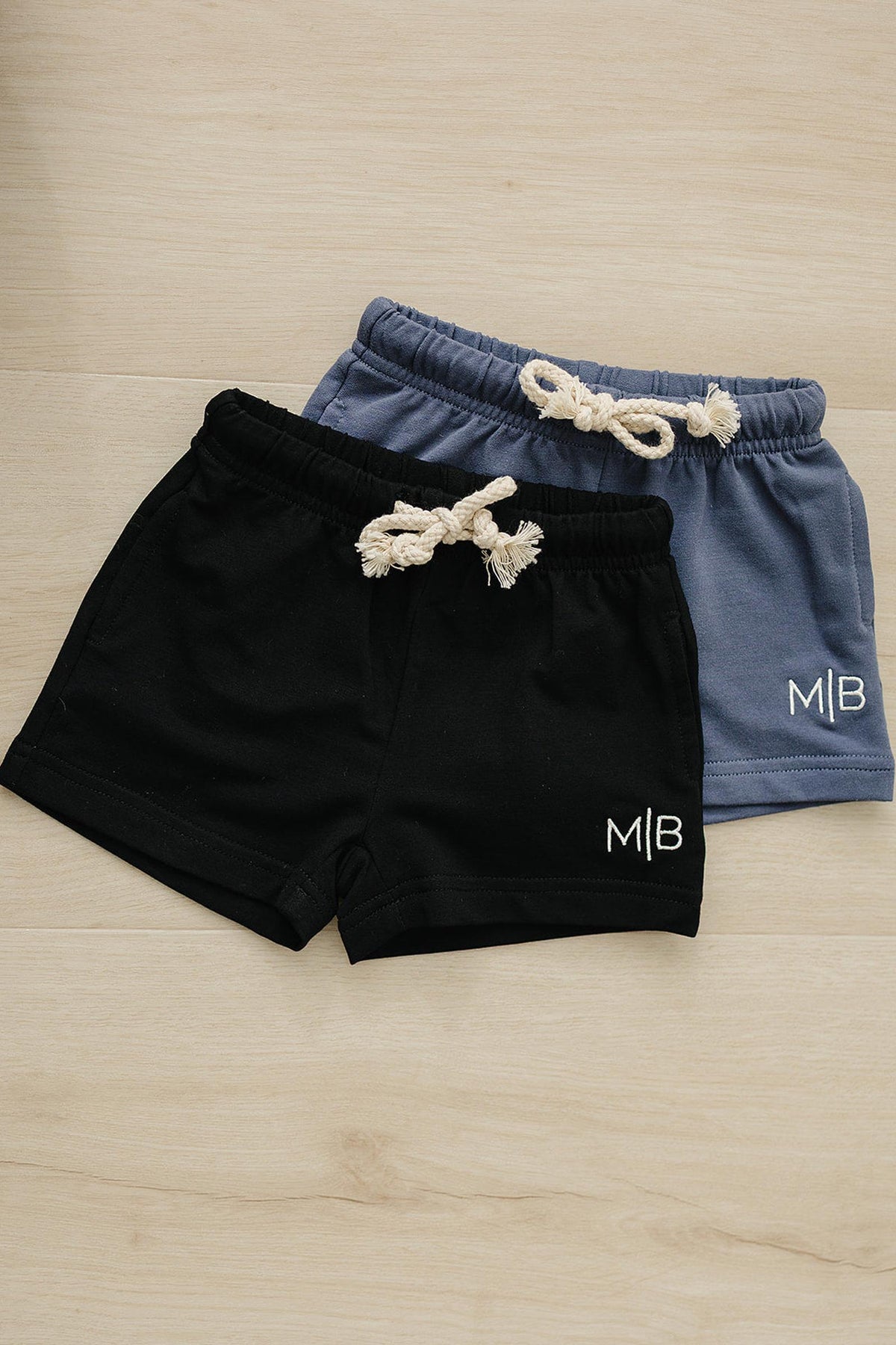 Two pairs of cotton shorts, one black and one blue, with rope drawstrings and M|B logo on wooden floor.