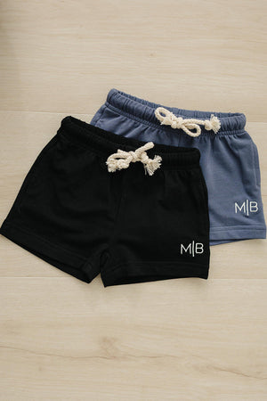 Two pairs of cotton shorts, one black and one blue, with rope drawstrings and M|B logo on wooden floor.