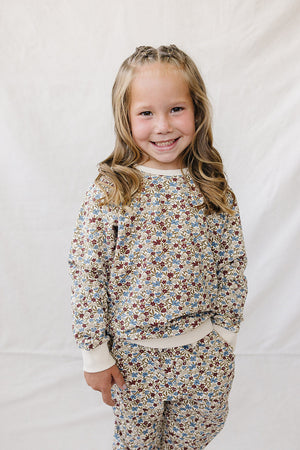 A smiling child wears a cozy, floral-patterned French terry cotton set featuring soft colors and a relaxed fit, ideal for comfortable play. The background is a simple, light fabric.