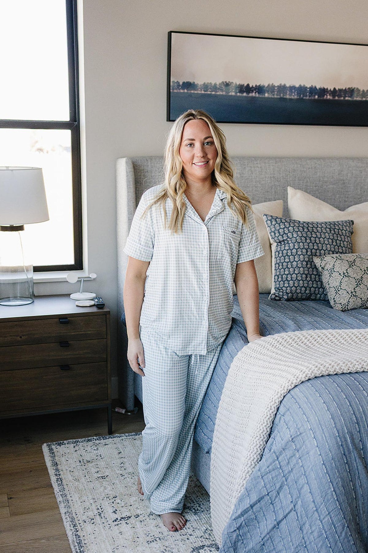 A woman in a light blue gingham pajama set stands by a bed with patterned bedding in a bedroom.