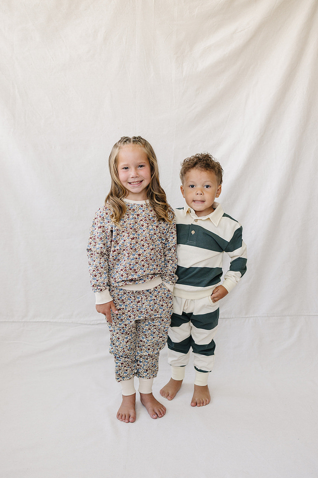 Two children stand side by side on a white backdrop. The girl wears a floral print outfit, while the boy is in a green striped collared set, both made from.