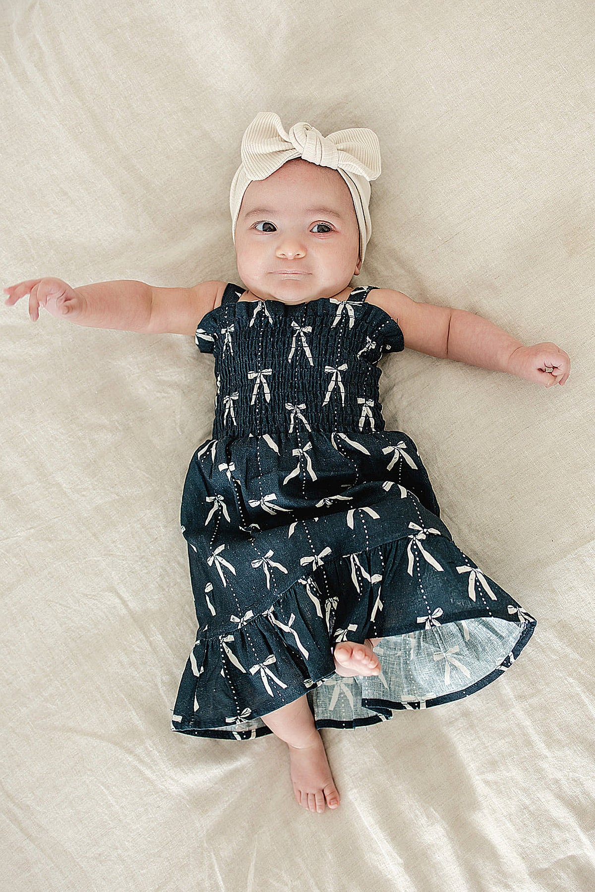 A baby wearing a navy dress with white bow prints and a cream headband lies on a beige blanket.