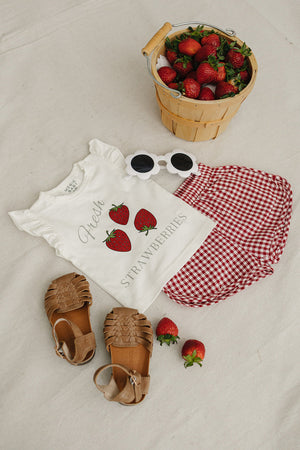 A playful white t-shirt features a Fresh Strawberries graphic, paired with a red gingham skirt, brown sandals, sunglasses, and a basket of fresh strawberries on a light background.