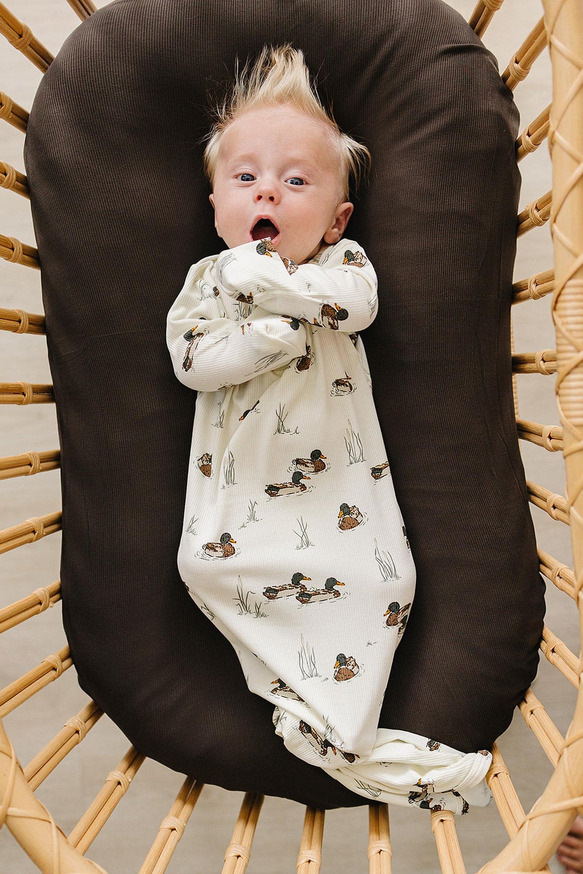 A baby with tousled hair lies in a cozy brown basket, wearing a soft, cream-colored gown featuring a playful duck print.