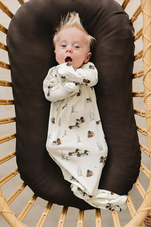 A baby with tousled hair lies in a cozy brown basket, wearing a soft, cream-colored gown featuring a playful duck print.