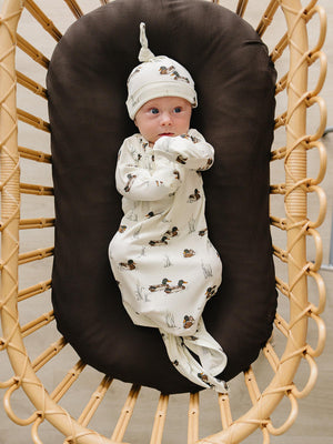 A newborn lies comfortably in a cozy, brown bassinet, wearing a soft, ivory knot hat and a matching swaddle, both featuring a whimsical duck print.