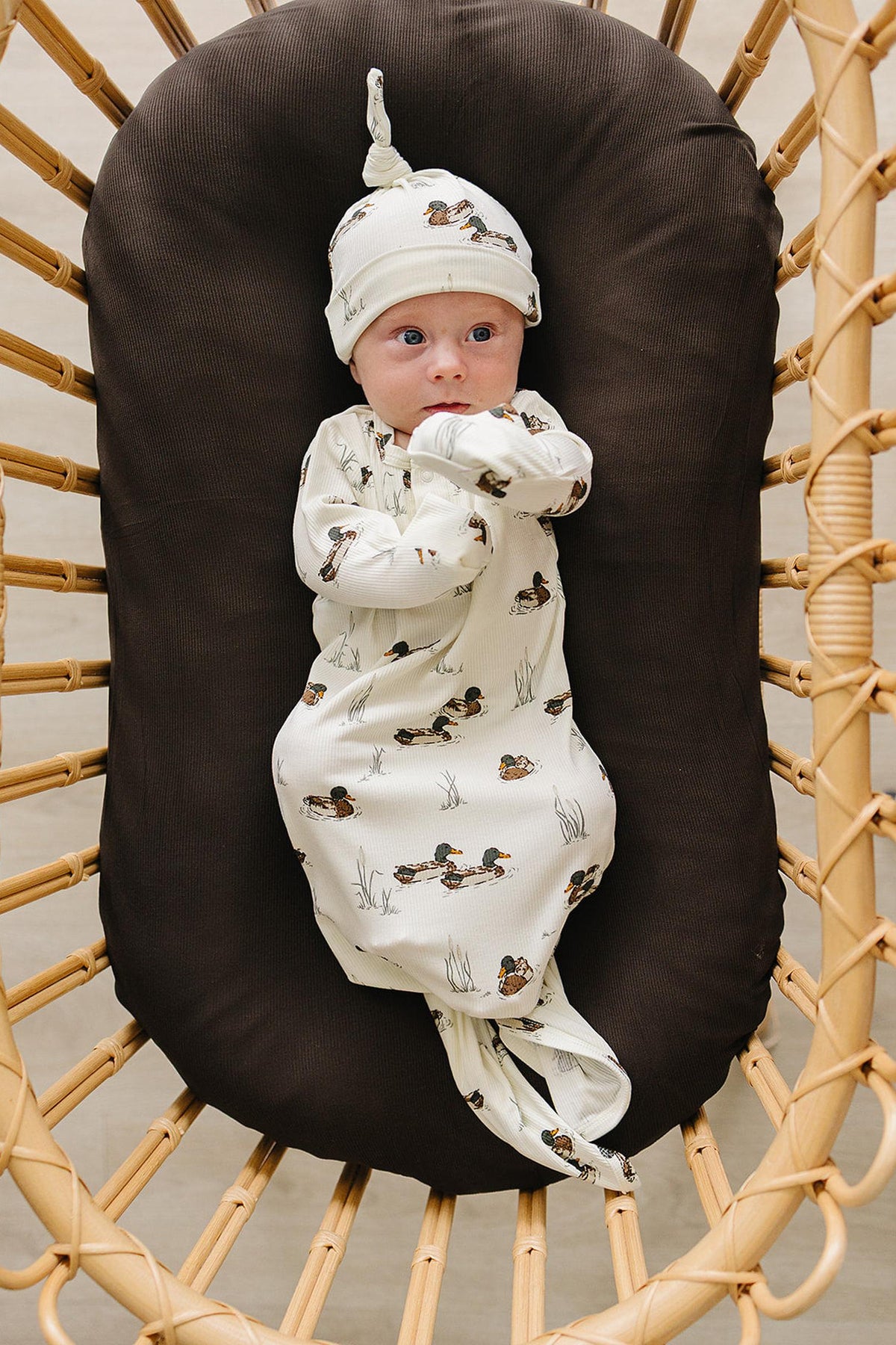 A newborn baby is swaddled in a cream-colored bamboo ribbed knot gown featuring a playful duck print, lying on a brown cushioned surface in a cozy setting.