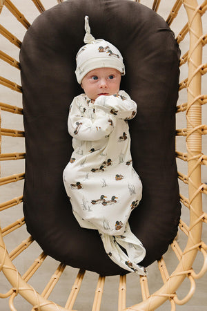 A newborn baby is swaddled in a cream-colored bamboo ribbed knot gown featuring a playful duck print, lying on a brown cushioned surface in a cozy setting.