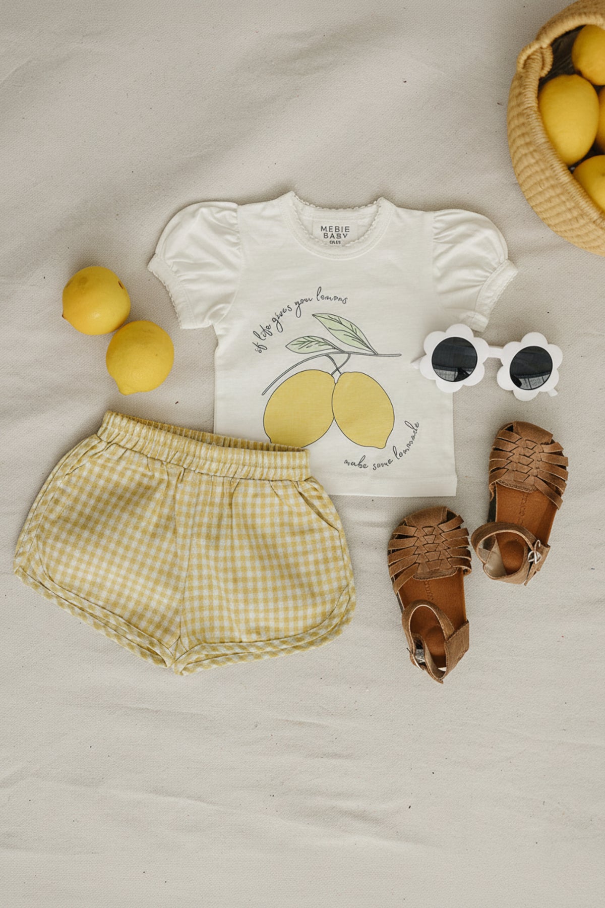 A white baby t-shirt with lemon graphic is paired with yellow gingham shorts, brown sandals, and white sunglasses, all arranged on a neutral background with lemons nearby.