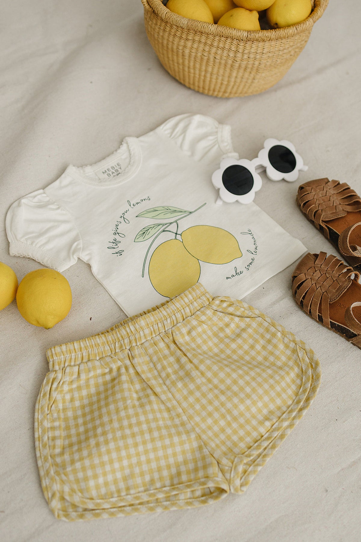 A yellow gingham shorts pair is displayed alongside a lemon-themed shirt, sunglasses, lemons, and brown sandals on a neutral fabric.