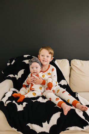 Two children in pumpkin-themed pajamas sit on a cream couch with a black and white blanket.