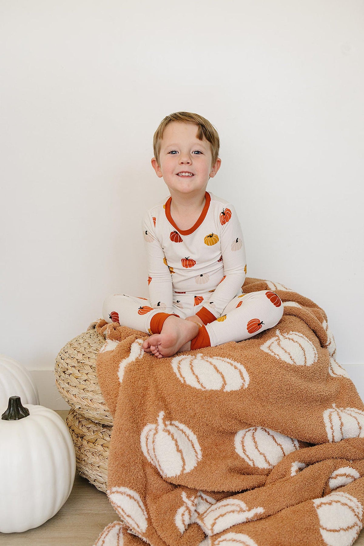 Child in a pumpkin-patterned pajama set sits on a pumpkin-themed blanket beside a white pumpkin.