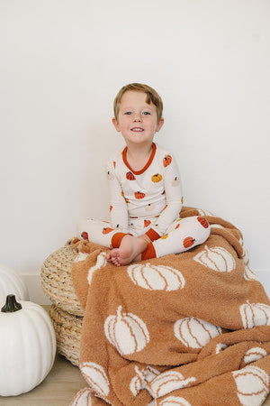 Child in a pumpkin-patterned pajama set sits on a pumpkin-themed blanket beside a white pumpkin.
