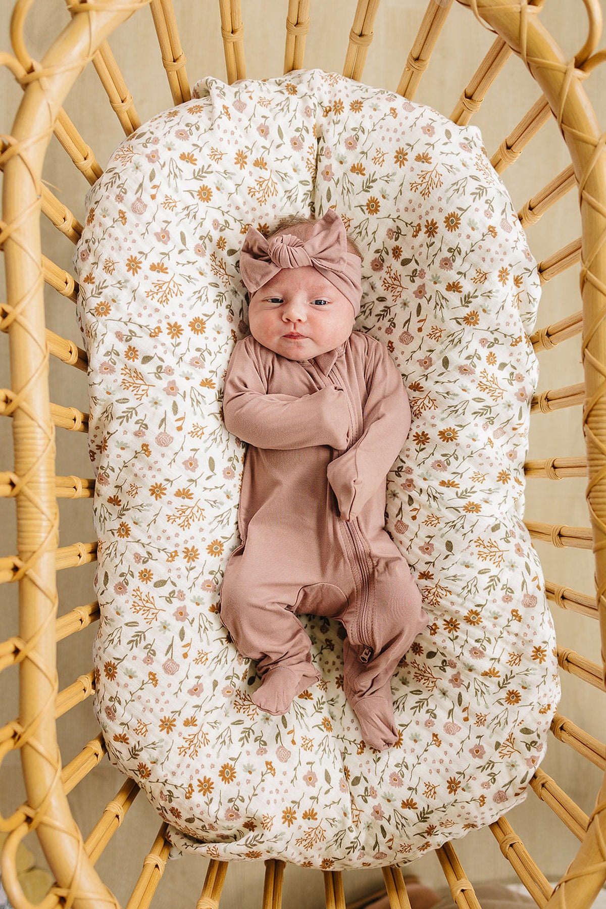 A baby in a dusty rose footed pajama and headband lies in a floral-patterned basket.