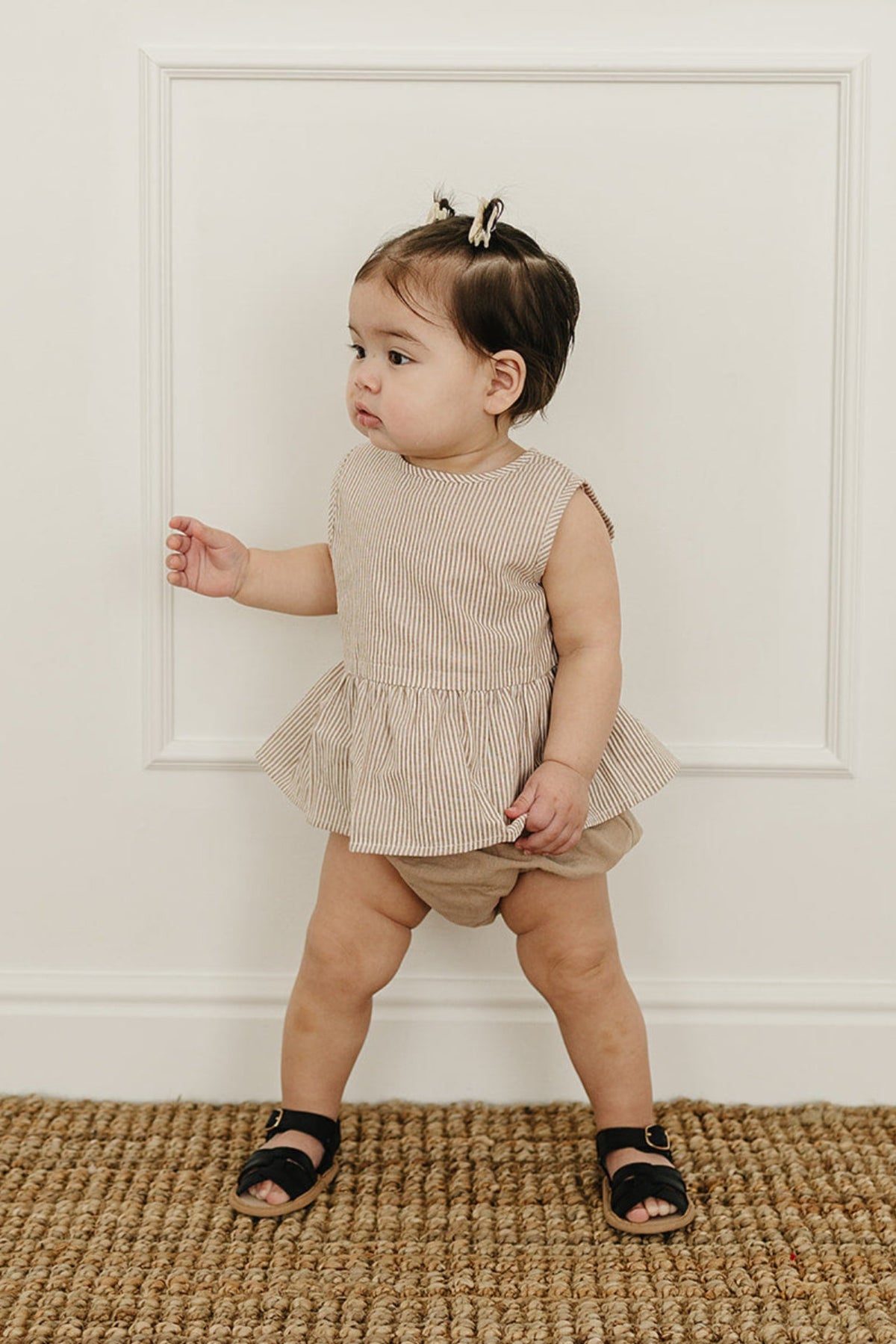 A toddler stands next to a white wall, wearing a tan striped peplum top and matching bloomers, with black sandals.