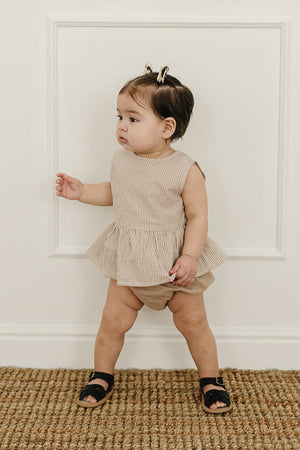 A toddler stands next to a white wall, wearing a tan striped peplum top and matching bloomers, with black sandals.