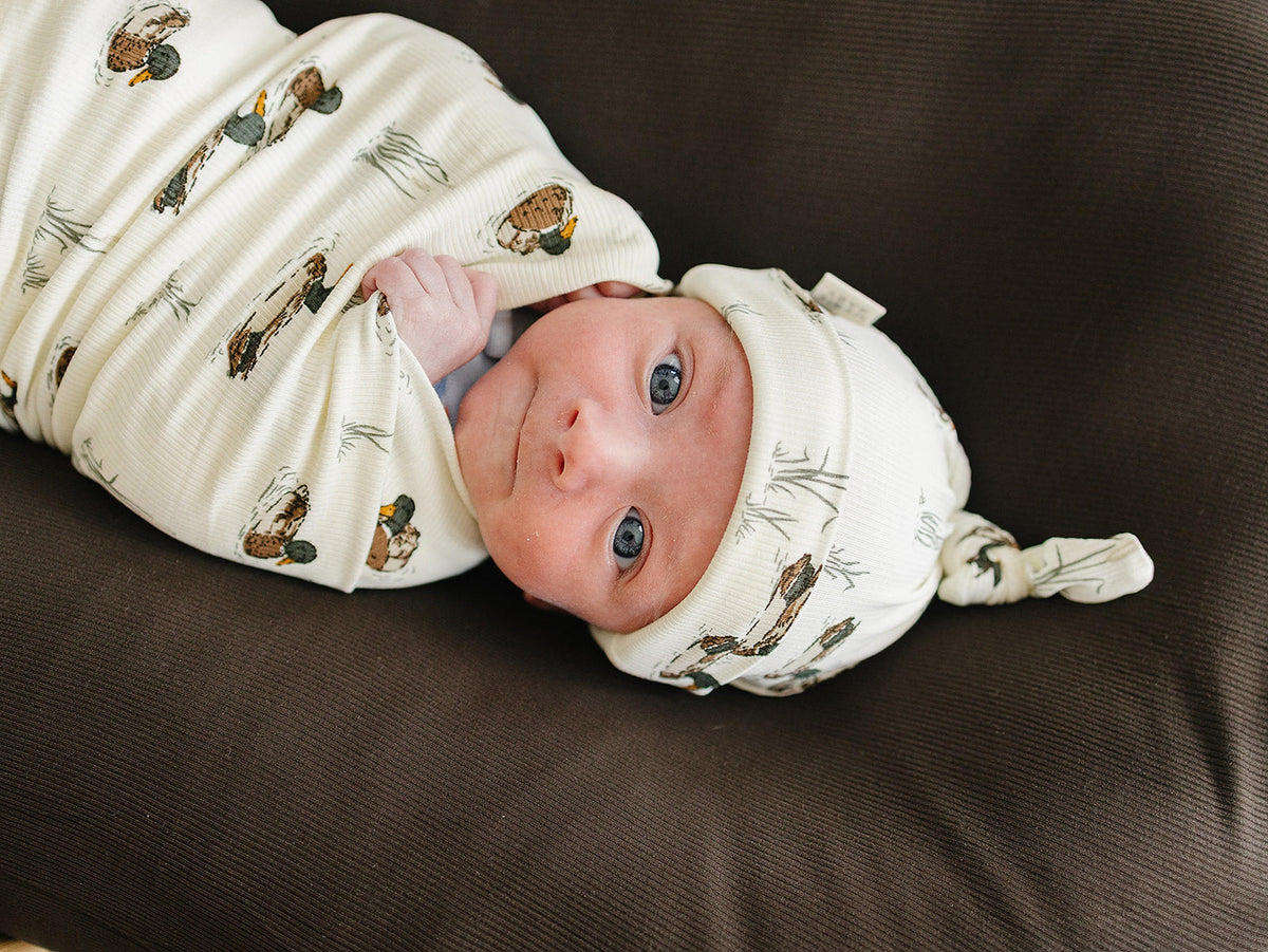 A newborn baby with blue eyes peeks out from a soft, ribbed bamboo swaddle featuring a duck print, wearing a matching knot hat.
