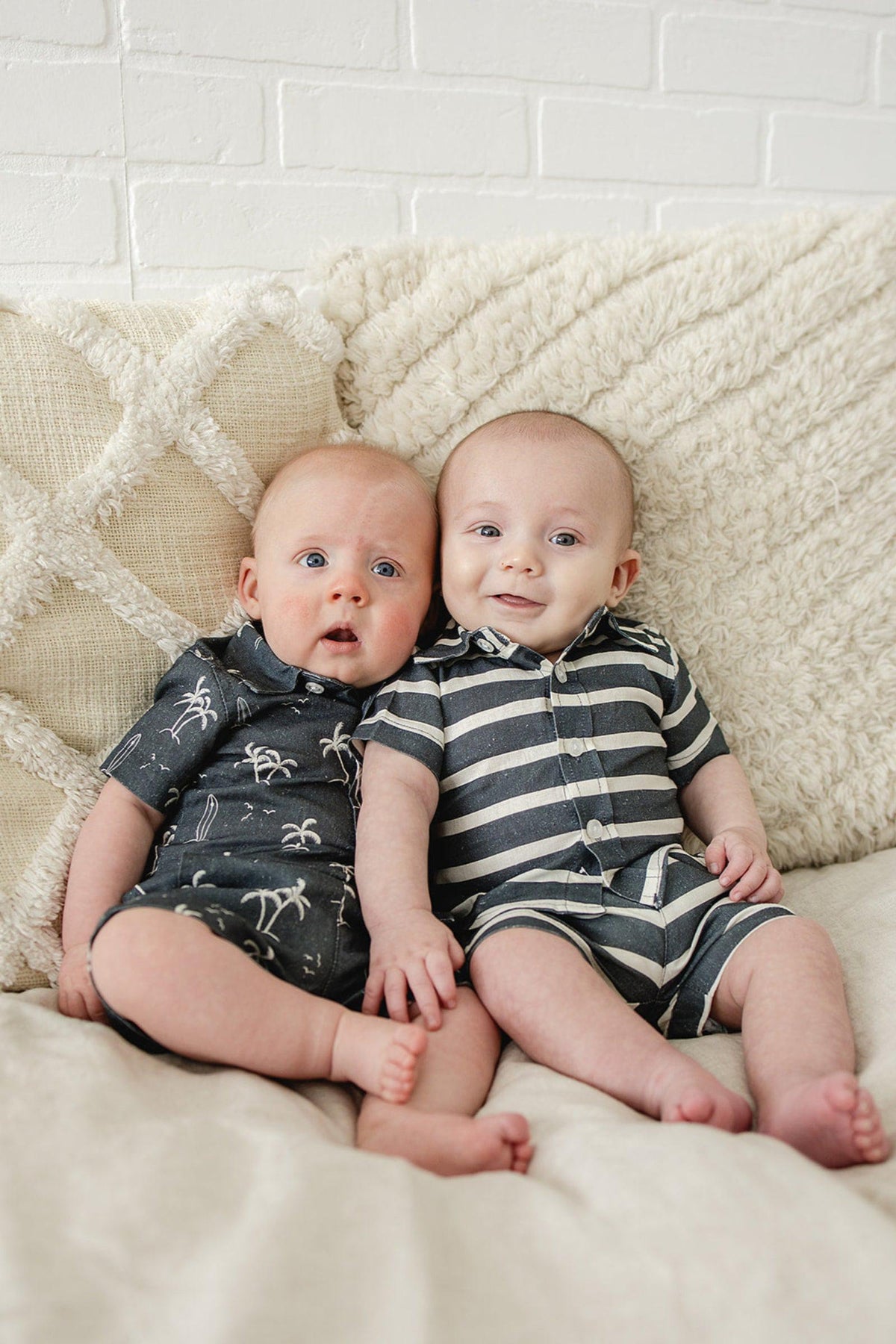Two babies wearing navy-themed outfits sit on a textured cream blanket against a white brick wall.