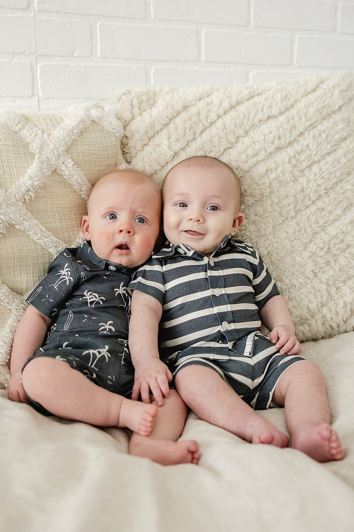 Two babies wearing navy-themed button-up rompers, one with stripes and the other with palm tree prints, sitting together.