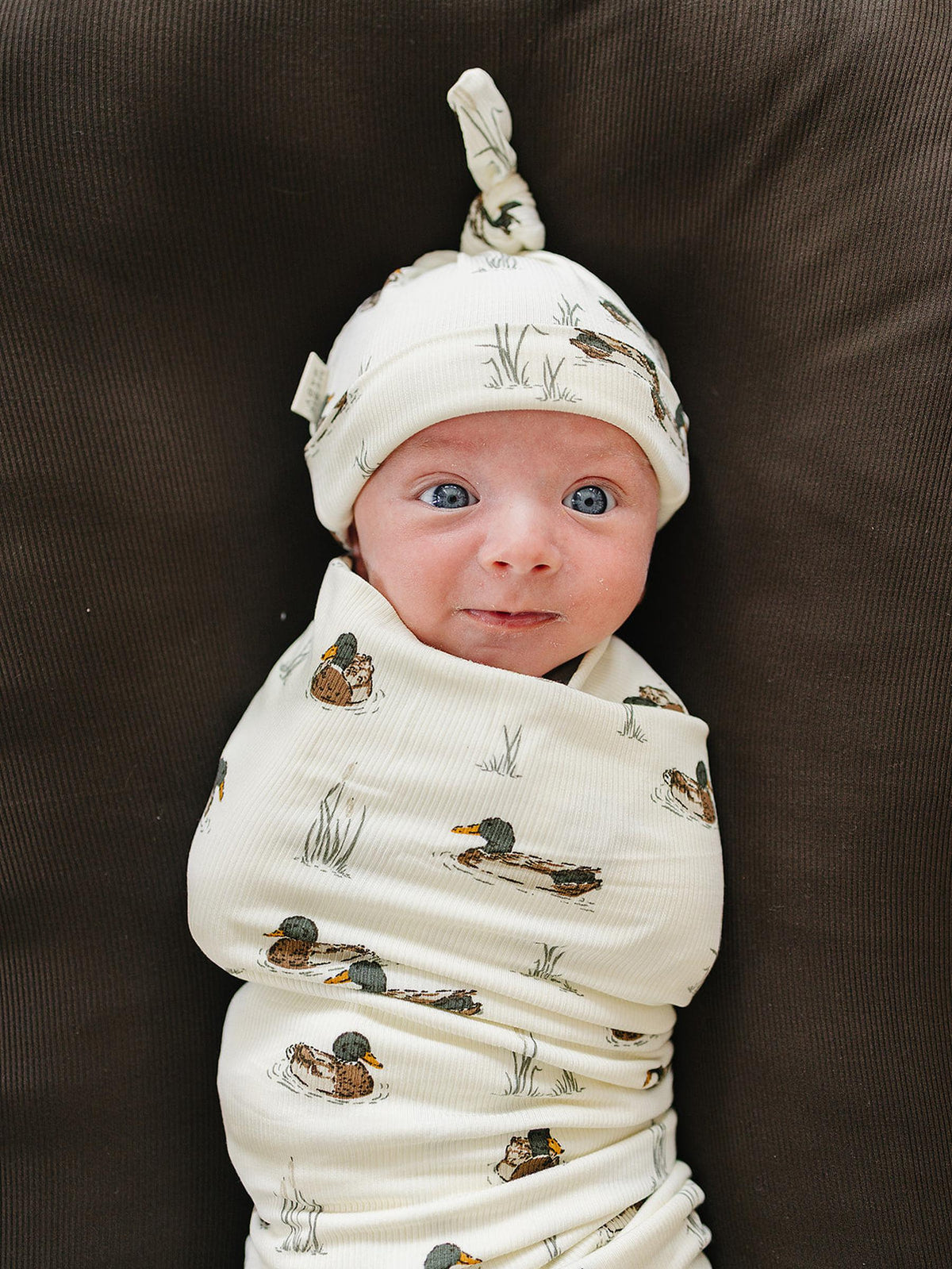 A newborn wrapped in a soft, ribbed bamboo swaddle features a duck print. The baby wears a matching knot hat, showcasing gentle colors and a cozy, snug fit.