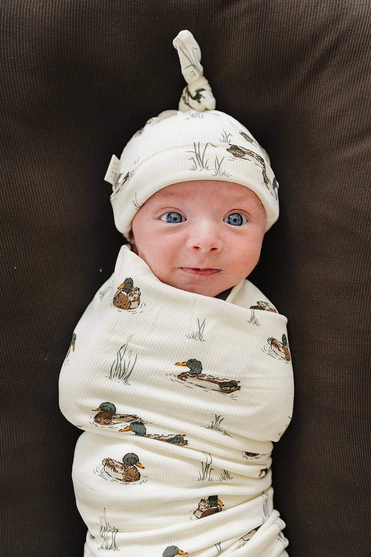 A smiling baby wrapped in a soft, ribbed bamboo swaddle featuring a duck print, complemented by a matching hat. The cozy fabric is gentle on delicate skin and eco-friendly.
