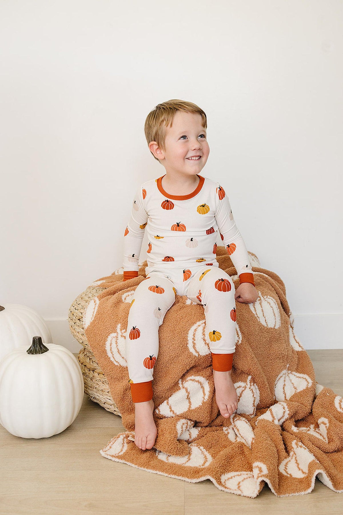 A child in pumpkin-patterned pajamas sits on a pumpkin-themed blanket, next to white decorative pumpkins.