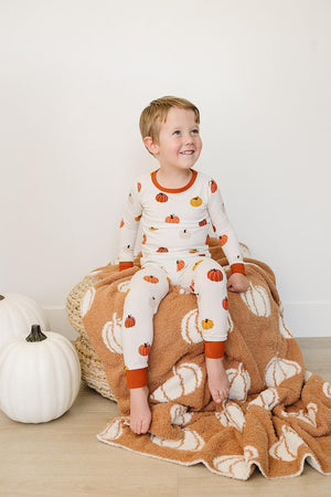 A child in pumpkin-patterned pajamas sits on a pumpkin-themed blanket, next to white decorative pumpkins.
