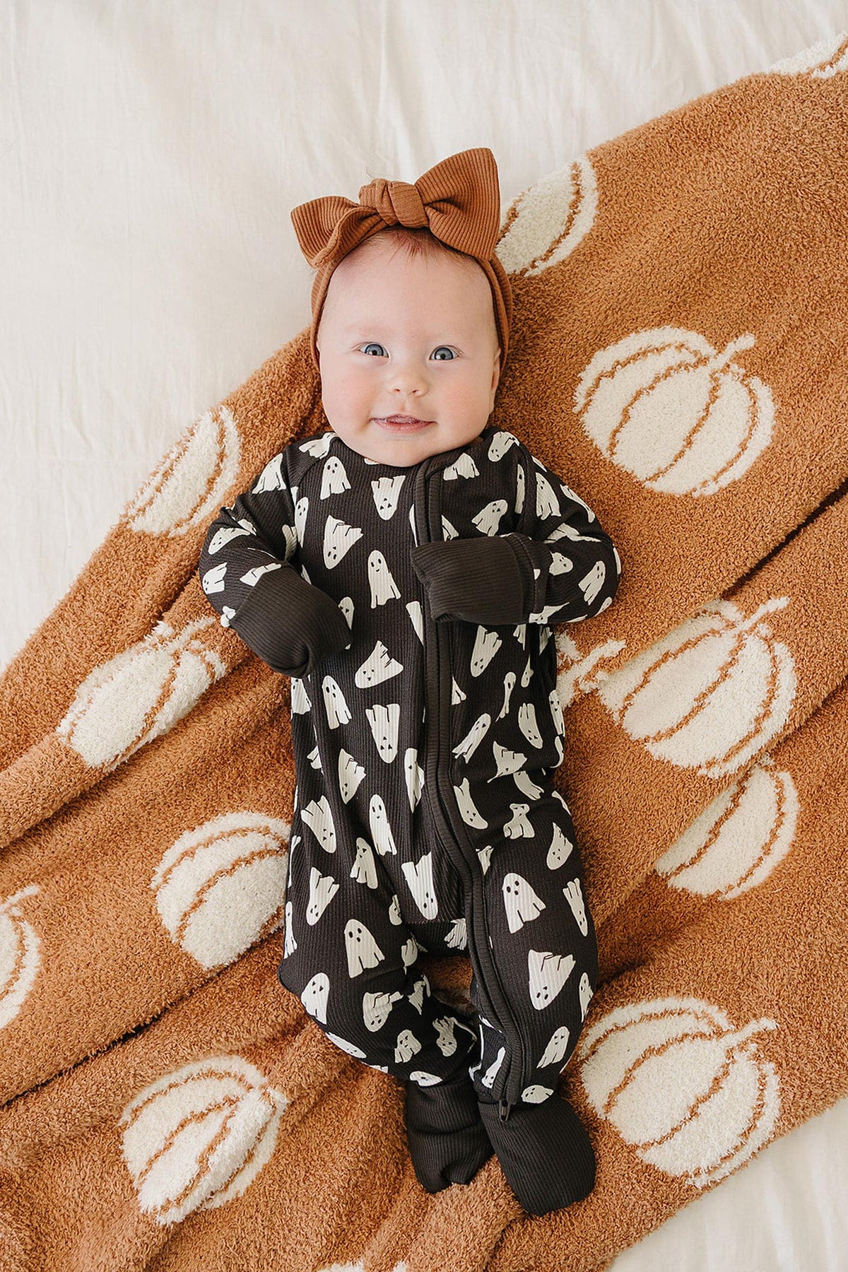 A baby in a black ghost-patterned pajama lies on a pumpkin-themed blanket, wearing a brown bow headband.