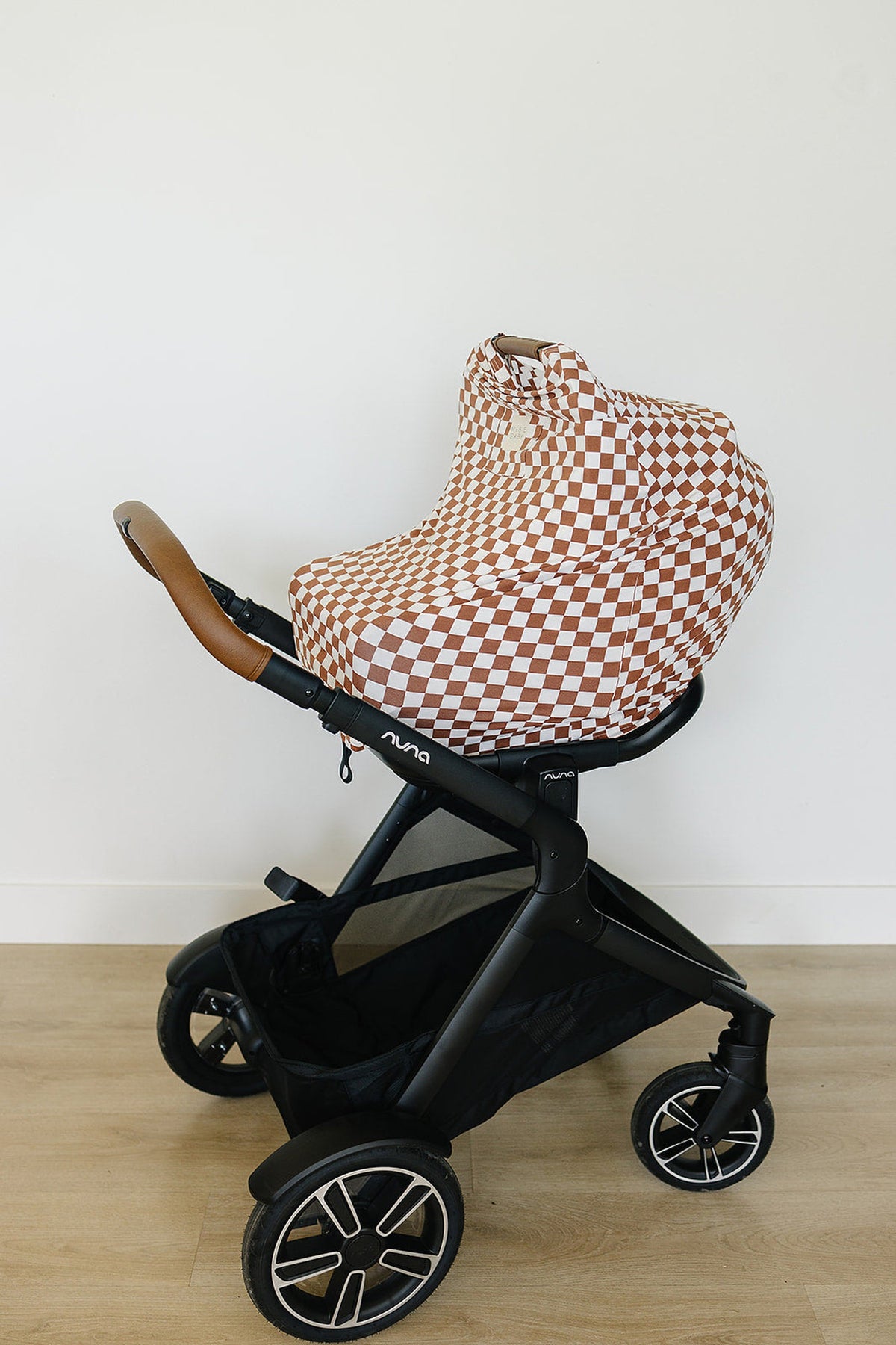 A stroller with a rust checkered bamboo cover over the car seat, featuring a black frame and leather handle.