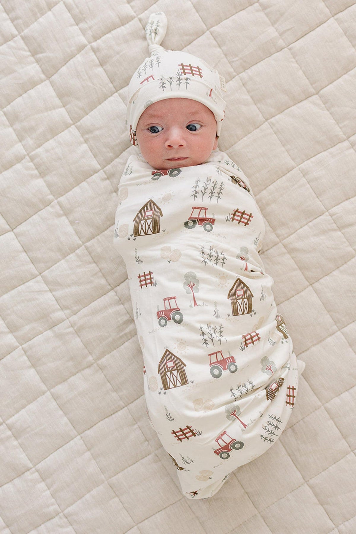A newborn baby, swaddled in a soft, cream-colored fabric featuring a charming farm print with tractors and barns, lies on a textured, light beige quilt.