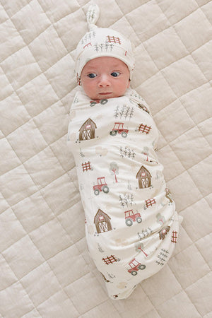 A newborn swaddled in a soft bamboo fabric features a charming farm-themed print with tractors and barns. The cozy swaddle is placed on a textured, neutral-colored quilted surface.