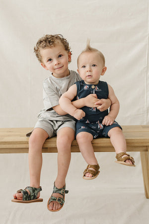 Two young boys sit on a wooden bench, one wearing a light grey short set and the other in a navy outfit featuring sailboats. Both look cheerful and comfortable.