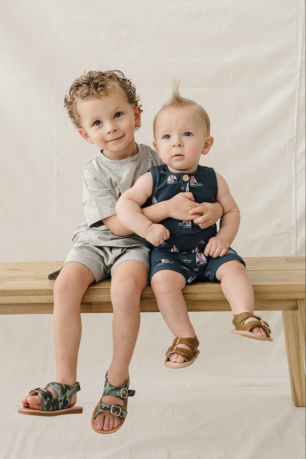 Two young boys sit on a wooden bench against a neutral backdrop. The older boy wears a gray shirt and shorts.