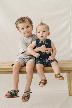 Two young boys sit on a wooden bench against a neutral backdrop. The older boy wears a gray shirt and shorts.