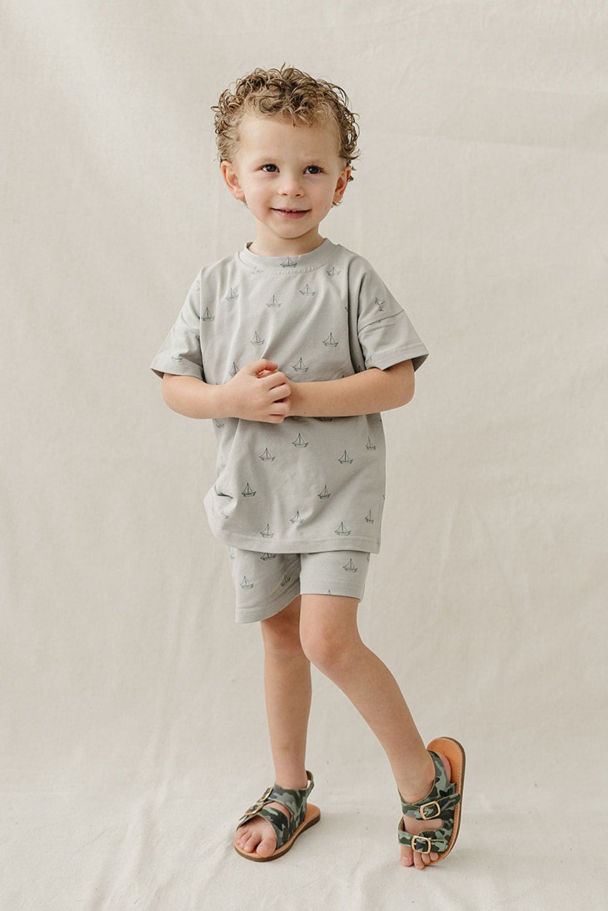 A young boy stands on a neutral background, wearing a light grey cotton shorts set featuring a sailboat print. He pairs the outfit with stylish sandals, exuding a playful vibe.