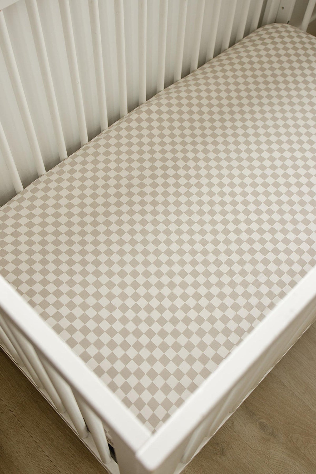 Taupe checkered crib sheet made from bamboo fabric on a white crib, placed on a wooden floor.