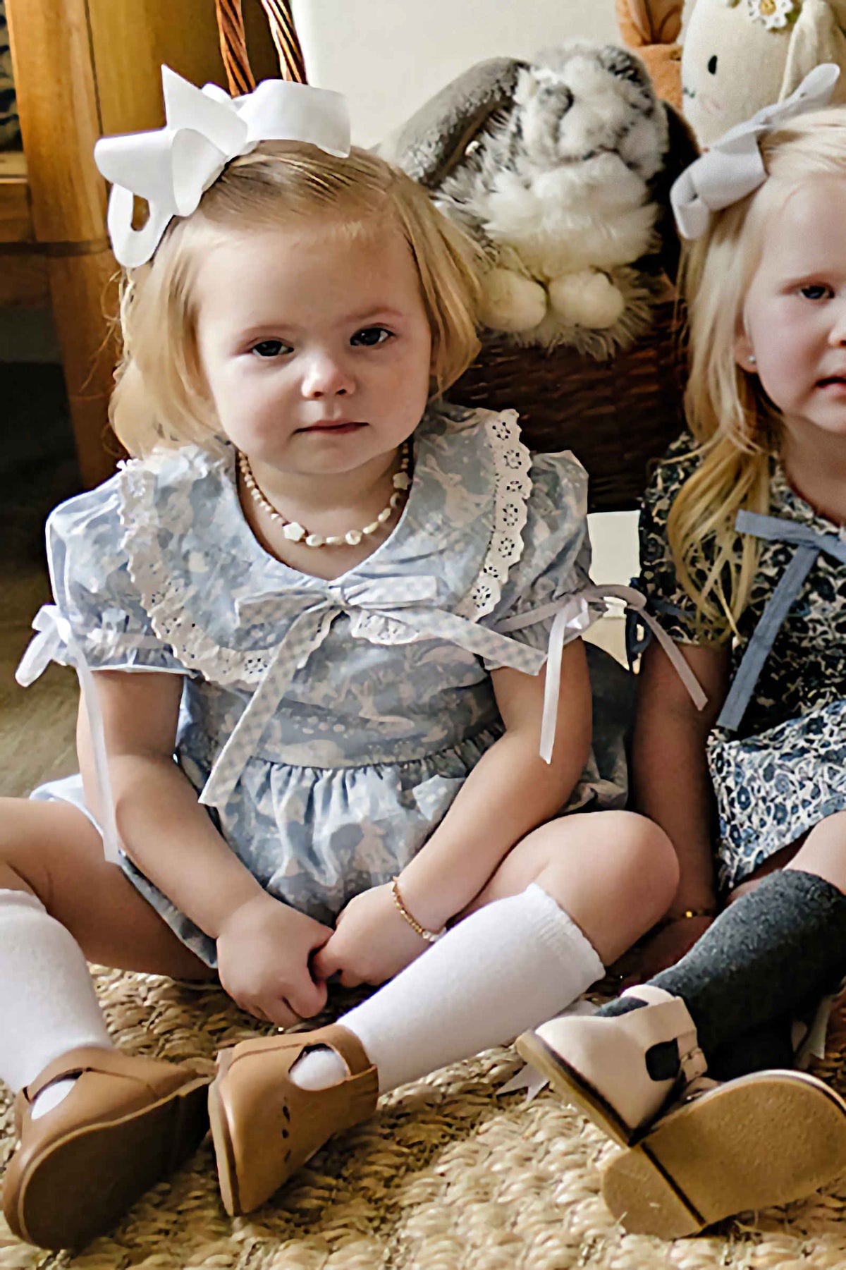 Two young girls sitting on a woven rug, wearing floral dresses with Peter Pan collars and white bows in their.